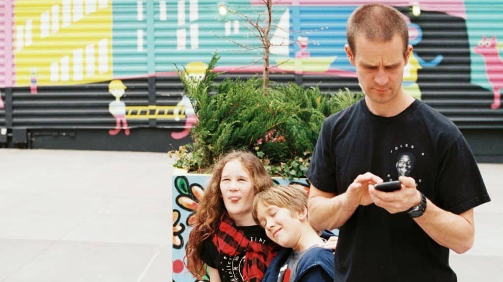 A man looks at a phone while two children pose playfully in front of him.