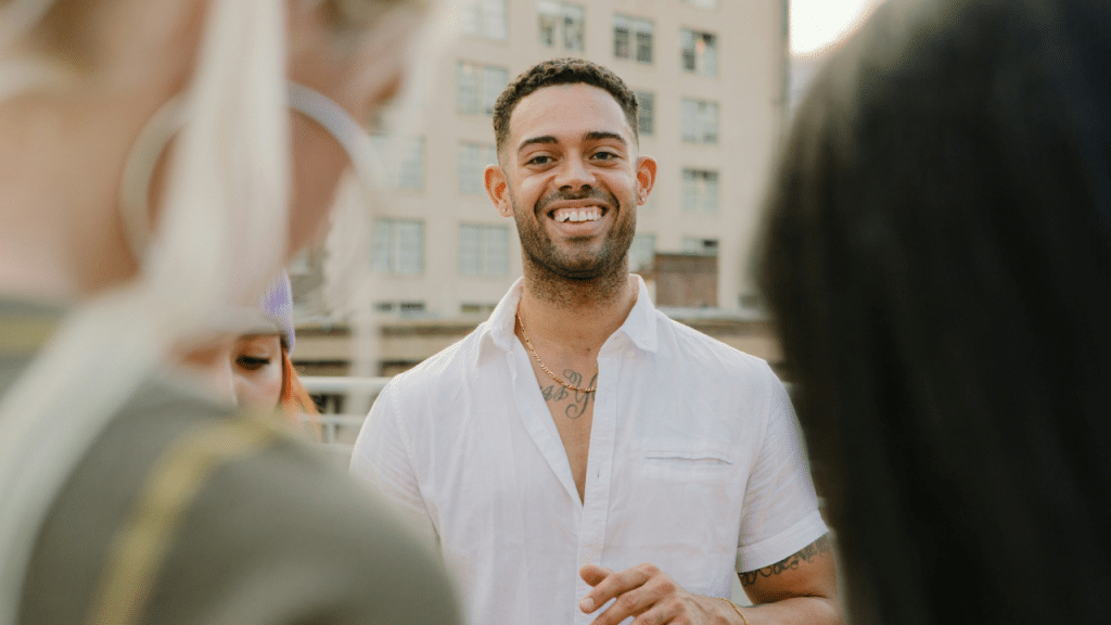 A smiling man in a white shirt stands outdoors while talking to a group of people.