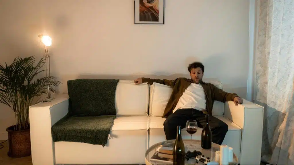 A man sitting on a couch with wine bottles and a glass on the table in front of him.