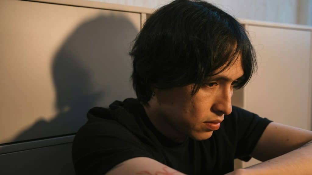 A man with dark hair looks downward with a somber expression while sitting indoors.
