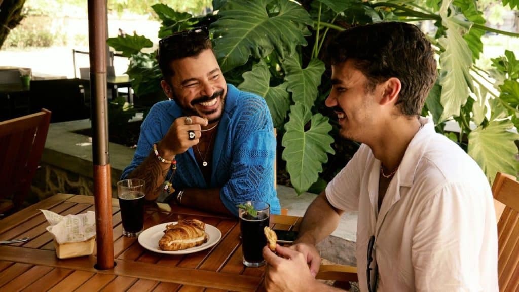 Two men sit at an outdoor table with food and drinks, laughing together near large green plants.