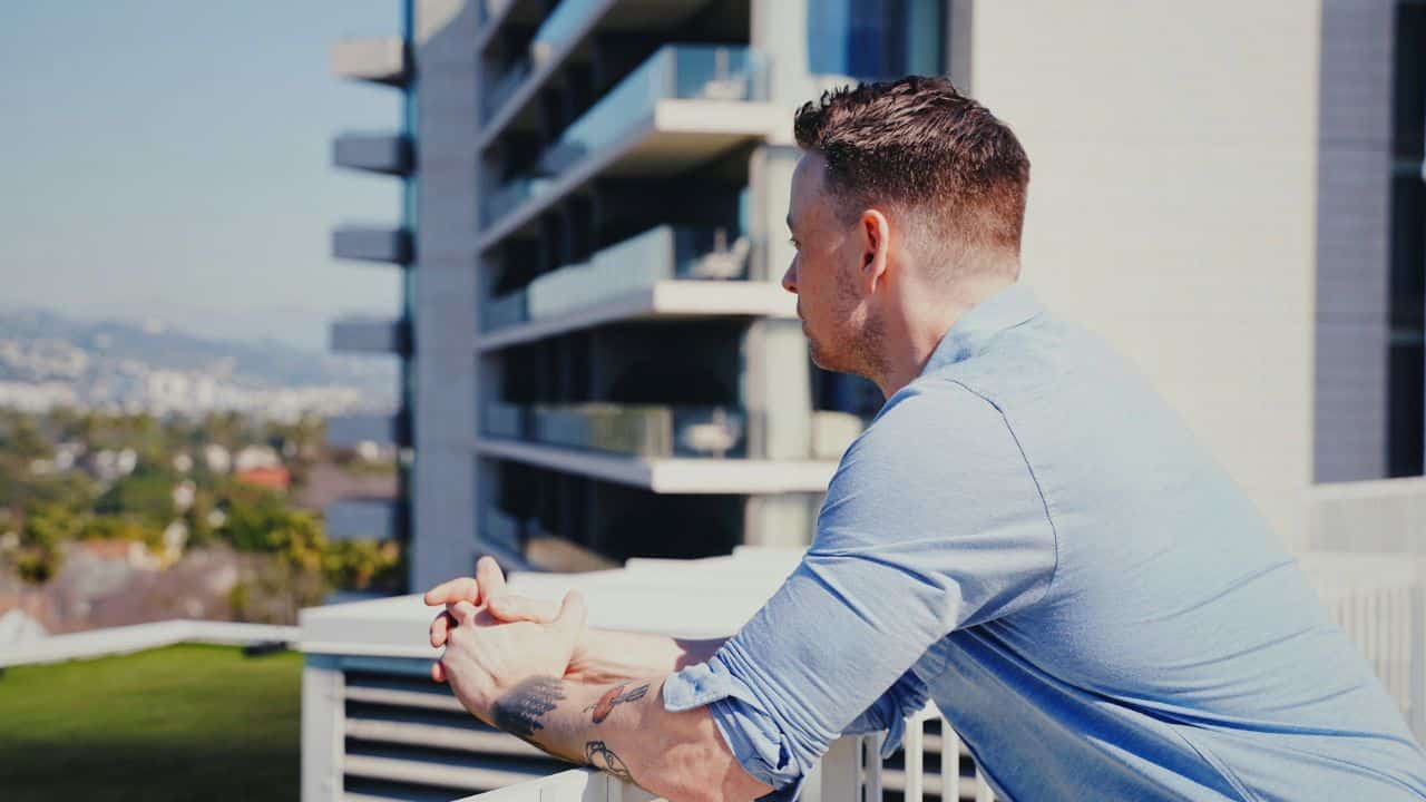A man with arm tattoos leans on a balcony railing looking at a city view.