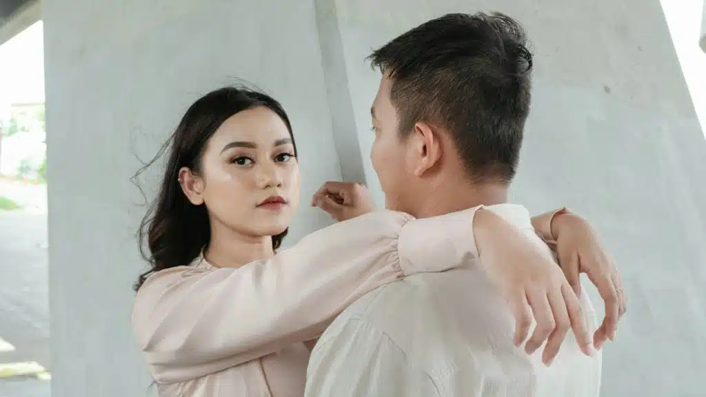 A woman looking at the camera while resting her arms on a man’s shoulders with an inscrutable expression.