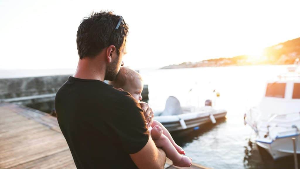 A dad holding a baby by a marina at sunset.