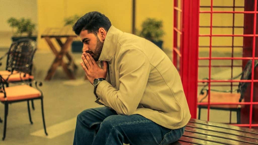 A man in a beige jacket sits on a bench with his hands pressed together.