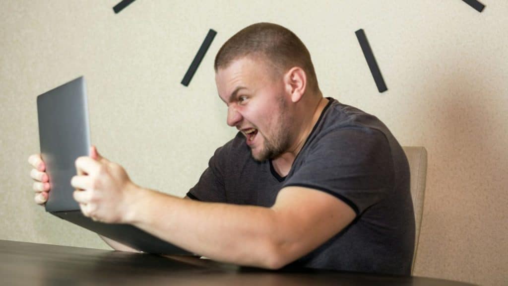 A man in a grey t-shirt shouts while holding a laptop with both hands.