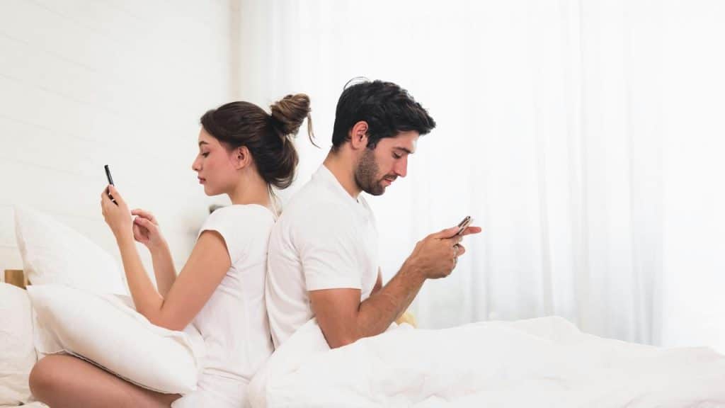 A couple sitting back to back in bed using their phones.