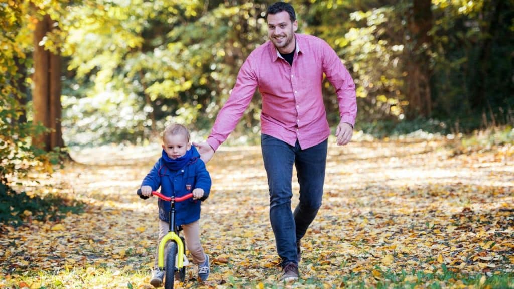 A man in a pink shirt walks behind a young child on a bicycle.