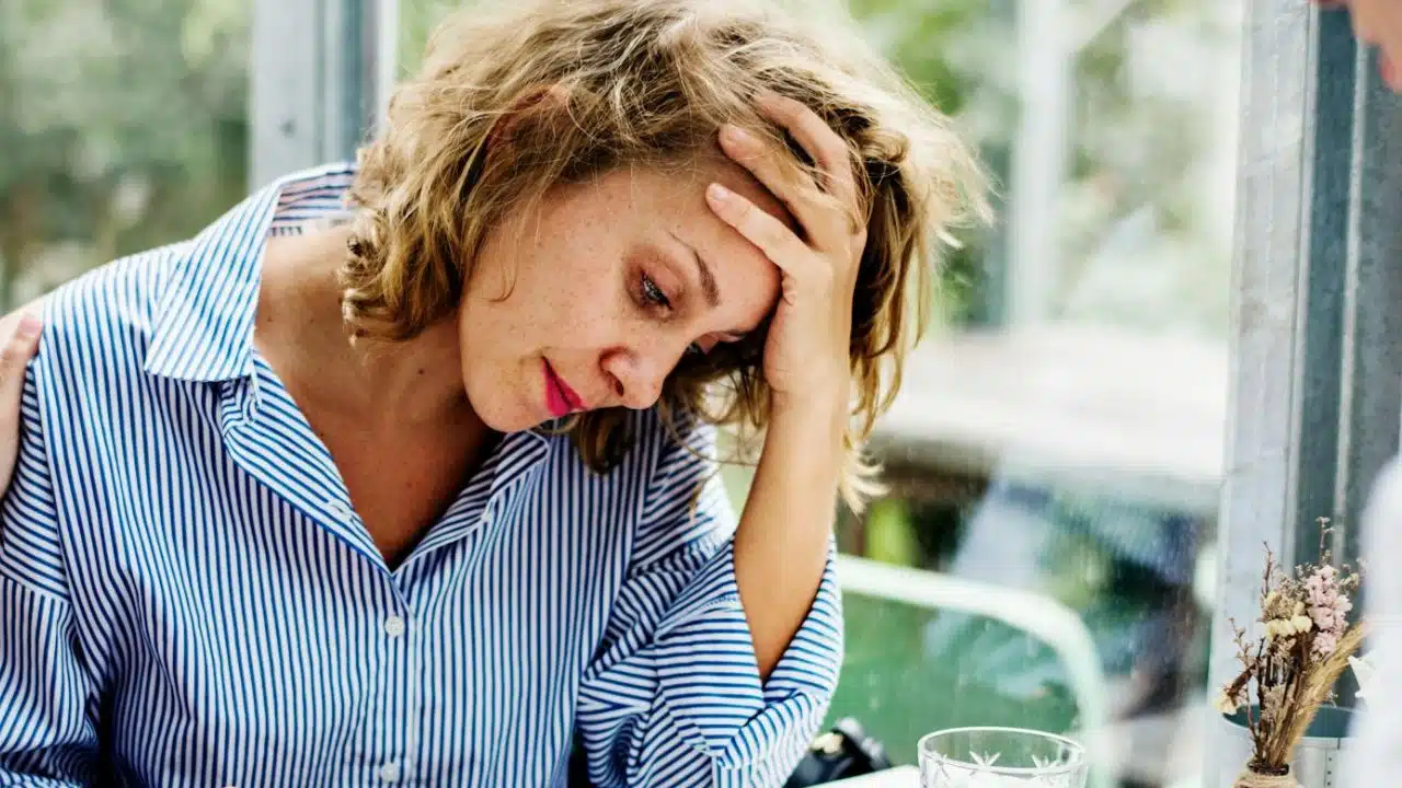 A woman with blonde hair rests her head in her hand with a downward gaze.
