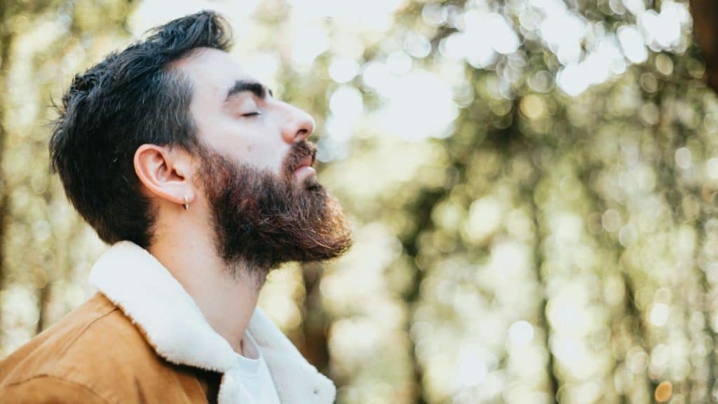 A bearded man in a sherpa-lined jacket closes his eyes against a soft, blurred forest background.