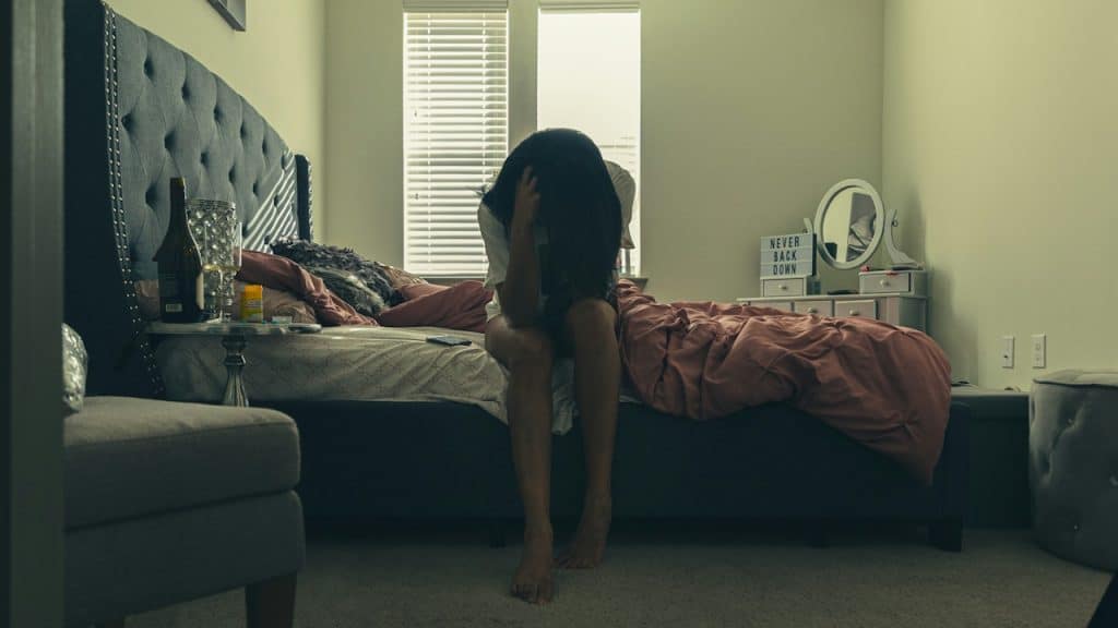 A woman sits on the edge of a bed in a messy room with her head in her hands.