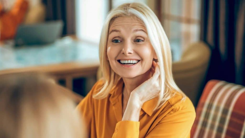 A blonde woman in an orange shirt smiles while looking at someone off-camera.
