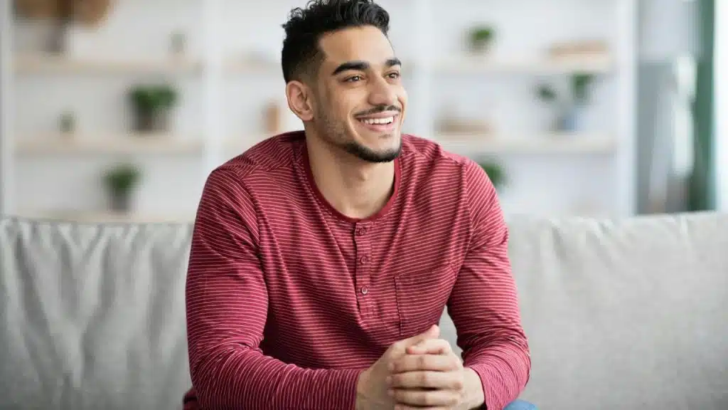 A smiling man with a beard sits on a gray couch wearing a red striped shirt.