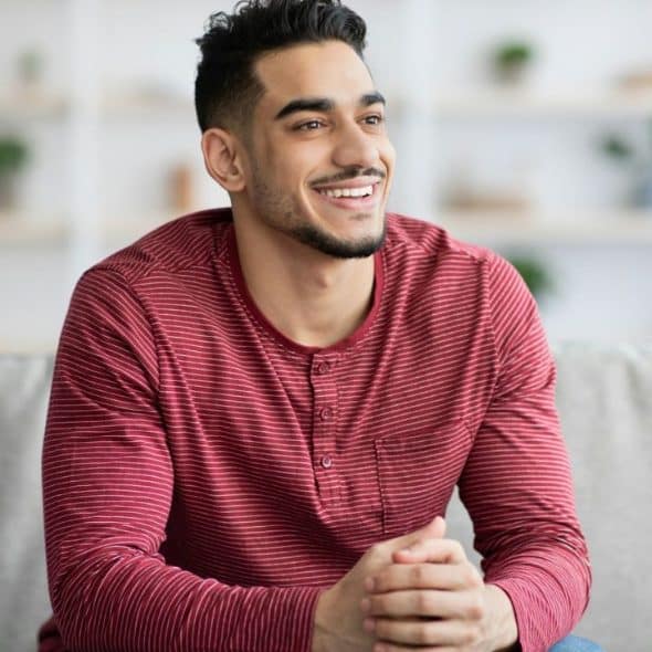 A smiling man with a beard sits on a gray couch wearing a red striped shirt.