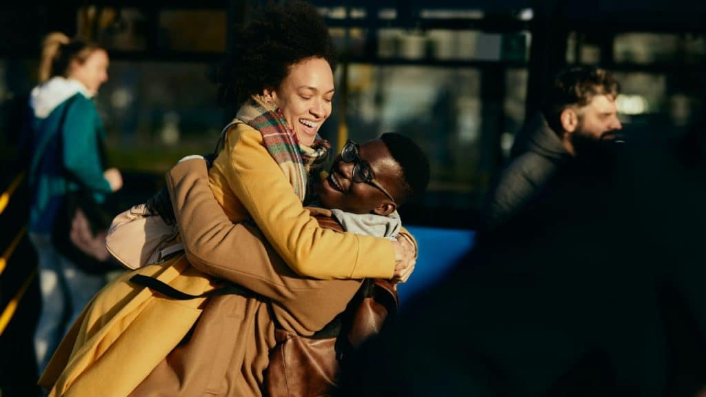 A laughing man and woman hug while standing outdoors in the sunlight.
