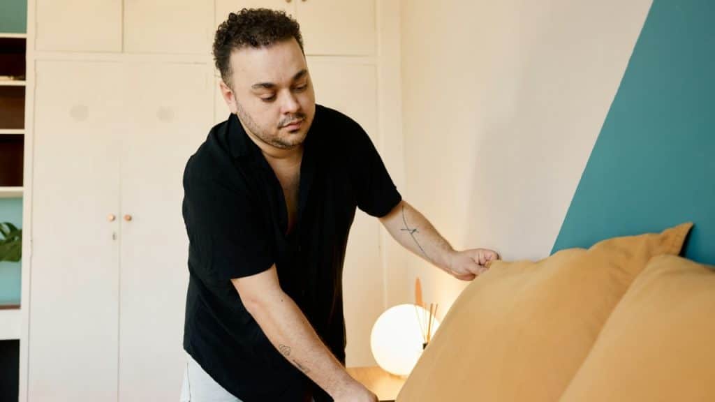 A man in a black shirt reaches down to adjust yellow pillows on a bed.