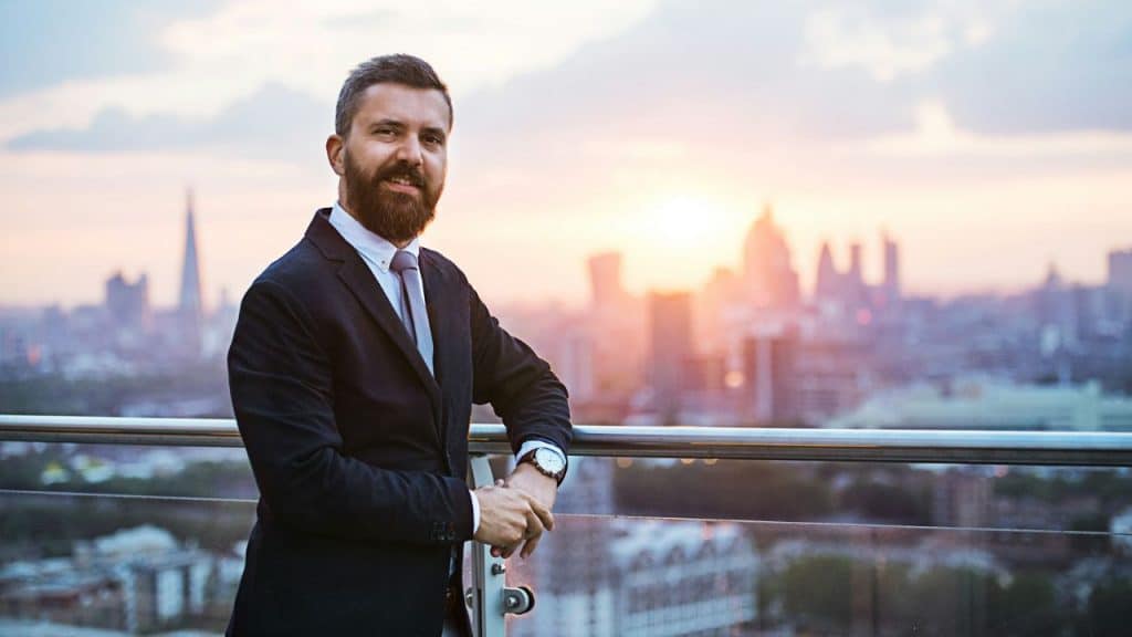 A bearded man in a suit stands on a balcony overlooking a city at sunset.