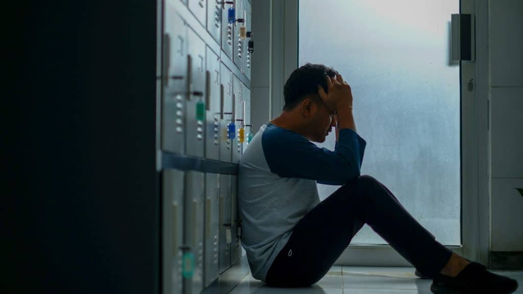 A man sitting on the floor while leaning against some lockers and holding his head in his hands.