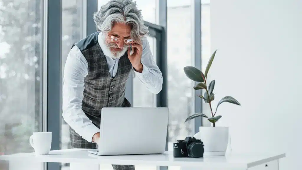 An older man wearing glasses using his laptop while talking on the phone.