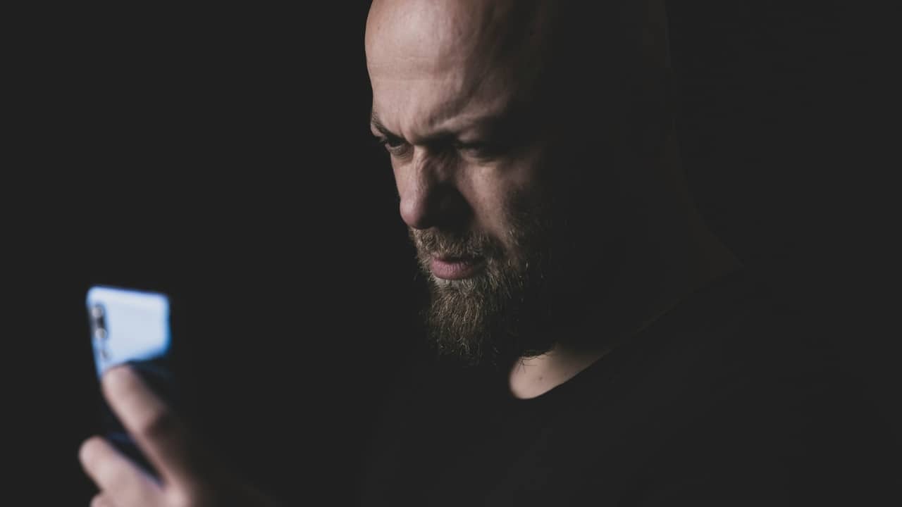 A bald man with a beard looks intently at a glowing smartphone screen in the dark.