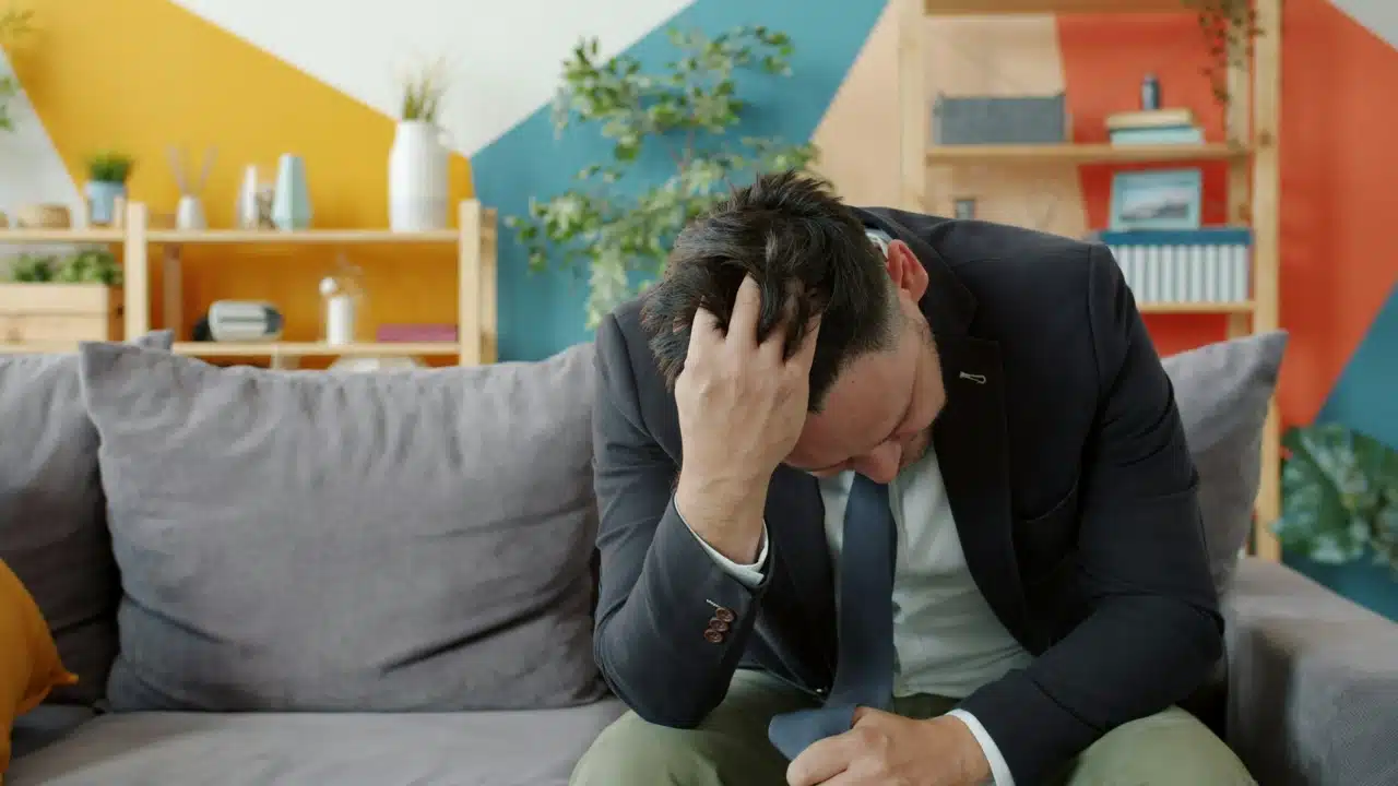 A man in a suit sits on a gray couch with his head in his hand.