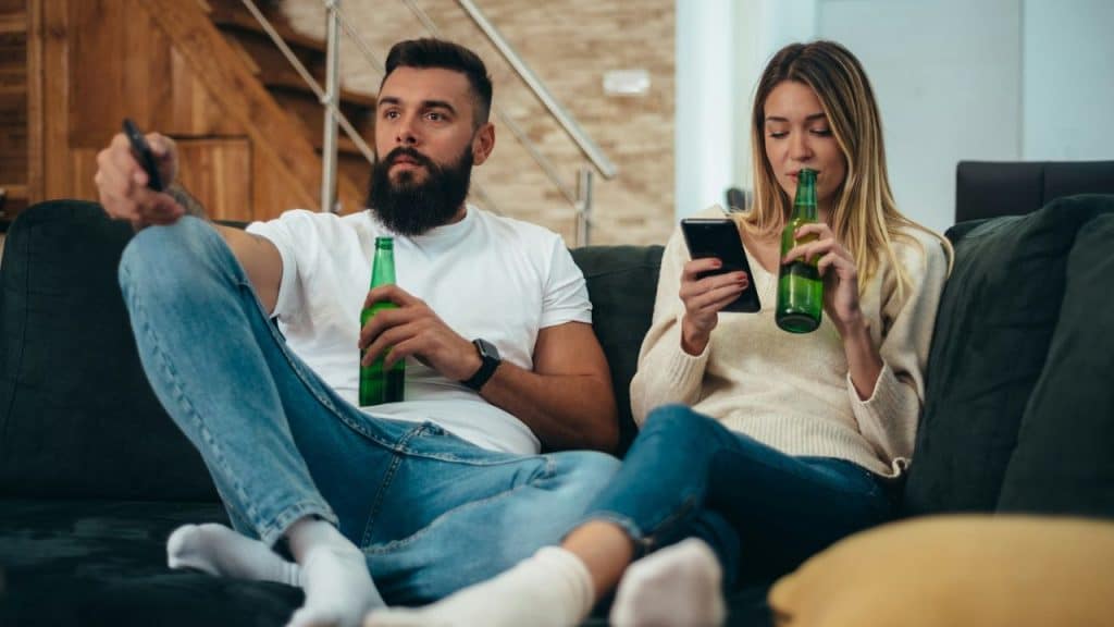 A man holds a remote and bottle while a woman beside him uses a smartphone.
