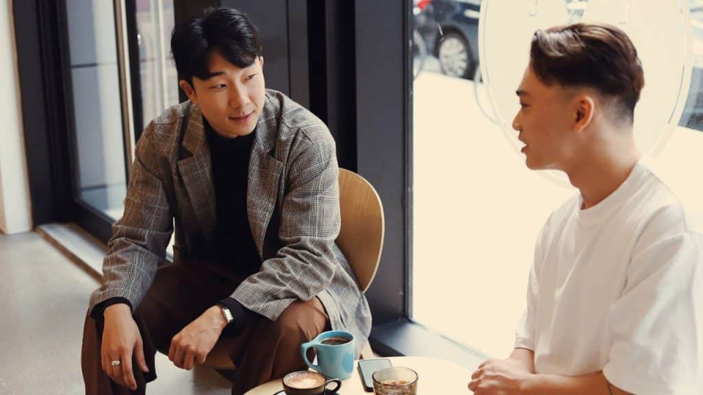 A man in a plaid blazer sits in a cafe, listening to another man speak.