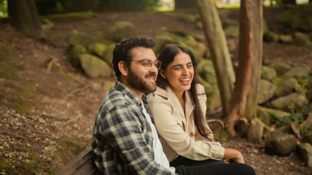 A bearded man wearing glasses sitting outdoors on a bench with a woman as they both smile.