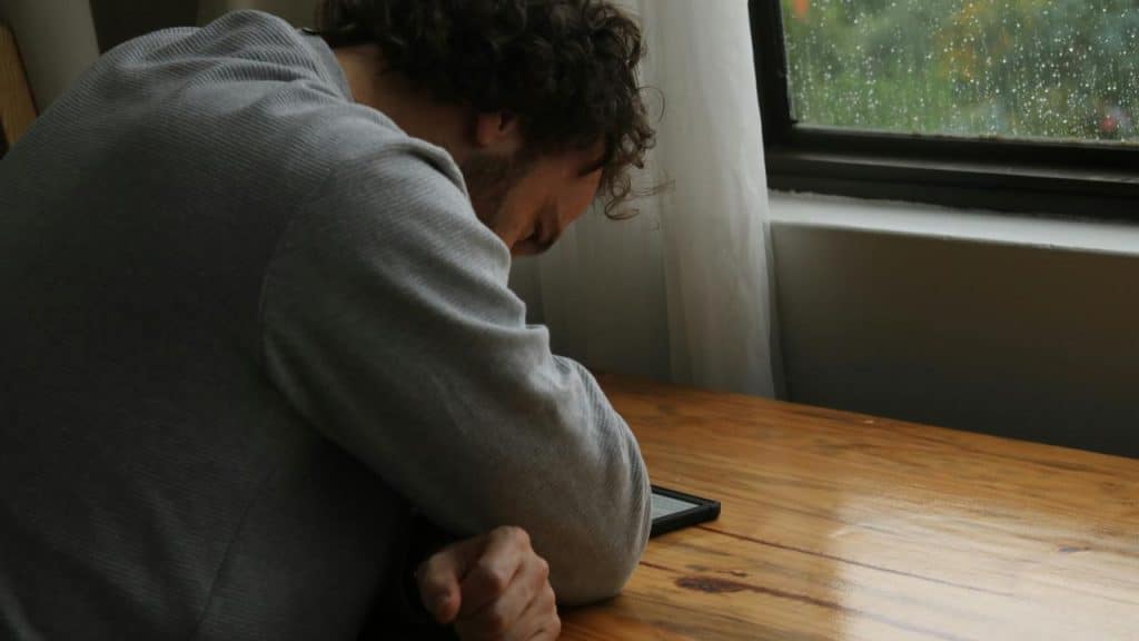 A person leaning over a table, looking down at a phone near a rain-streaked window.