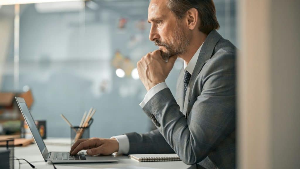 A man in a grey suit stares at a laptop screen with his hand on his chin.