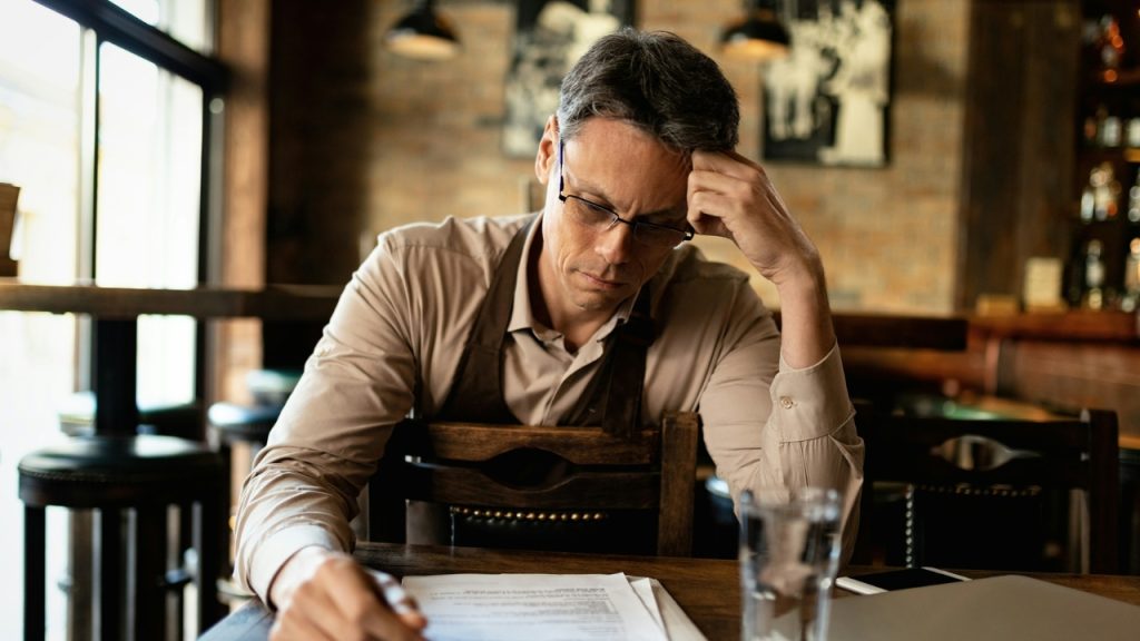 A man wearing glasses sits at a table looking down at papers with his hand on his head.