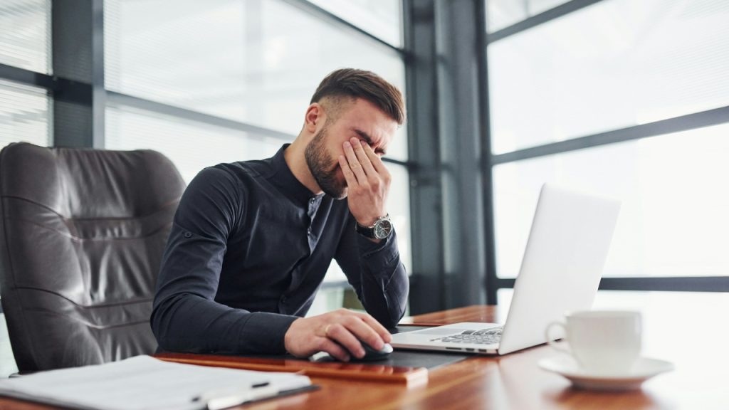 A man at a desk rubs his eyes while sitting in front of an open laptop.