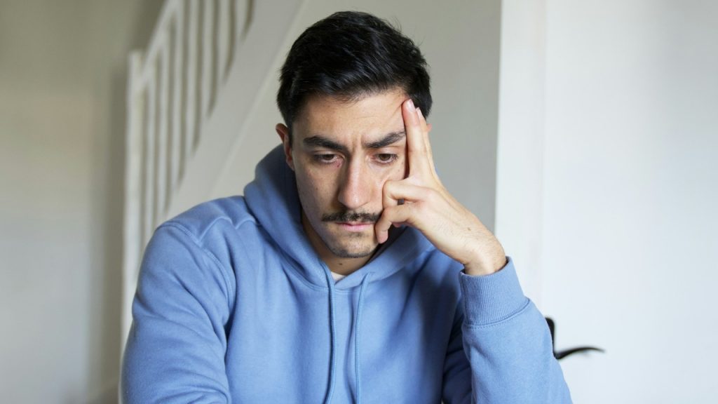 A man in a blue hoodie rests his head on his hand while looking down.