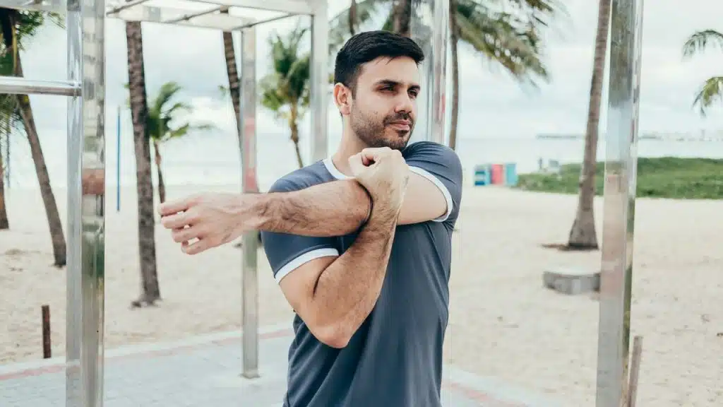 A man in a gray t-shirt stretches his arm across his chest on a beach.