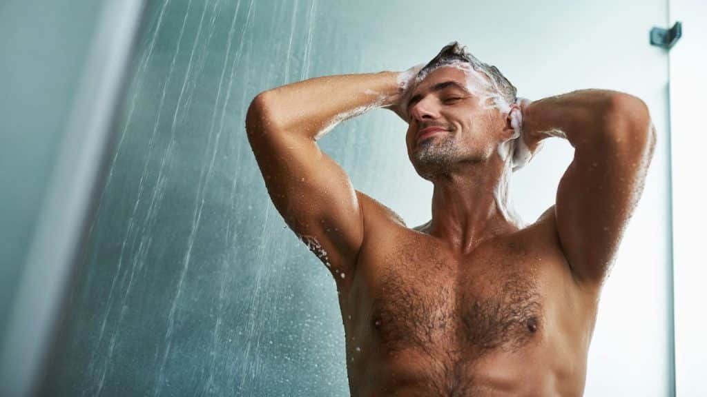 A shirtless man with his eyes closed washes his hair with soap in a shower.