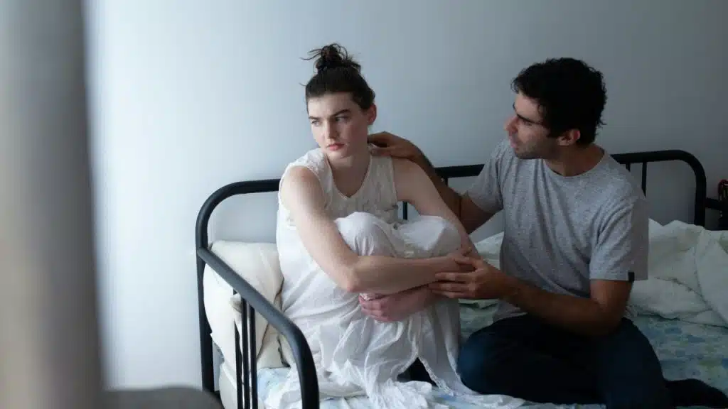 A man in a grey shirt sits on a bed and touches a woman's shoulder.