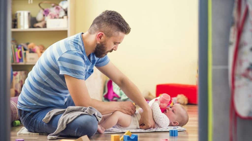A man feeding a baby with a bottle while changing their diaper.