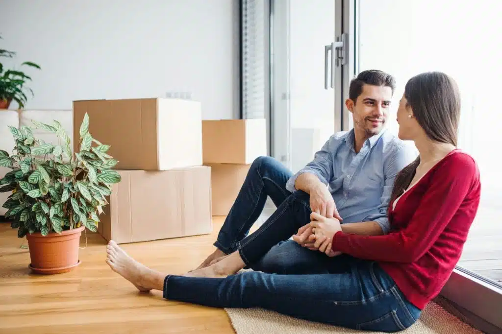 A man and woman talking while sitting on the floor and leaning against a window.