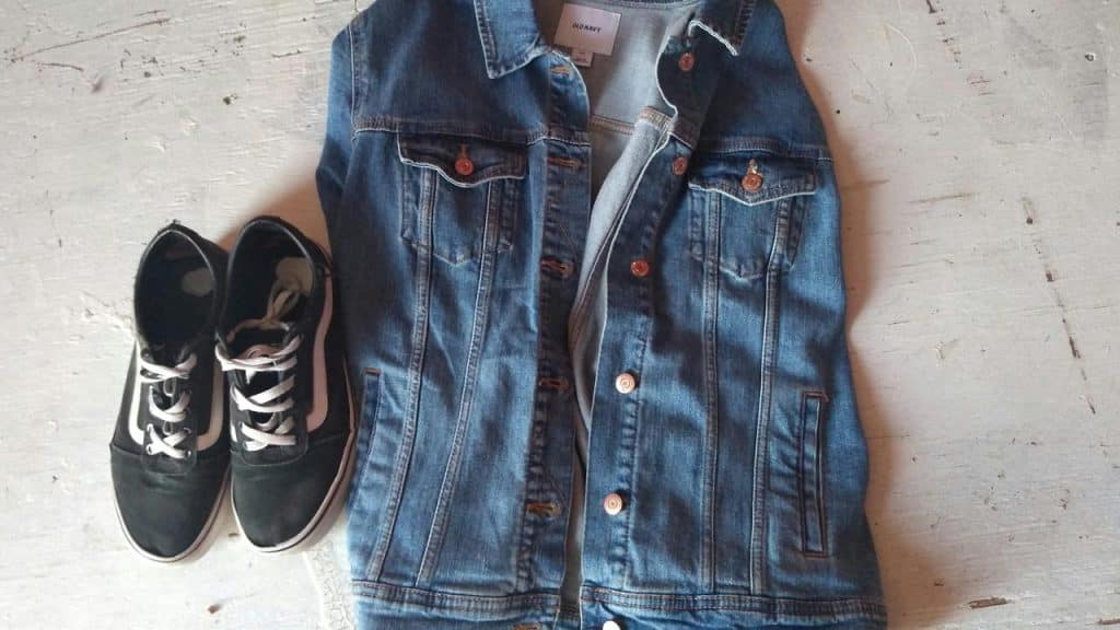 A denim jacket beside a pair of black sneakers on a light floor.
