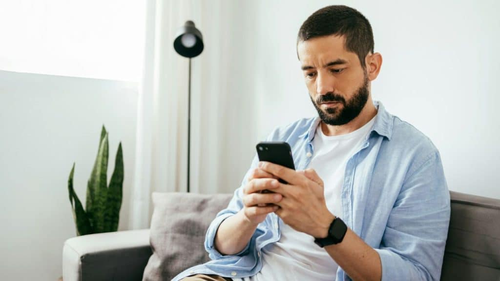 A bearded man in a light blue shirt sits on a sofa looking at his smartphone.