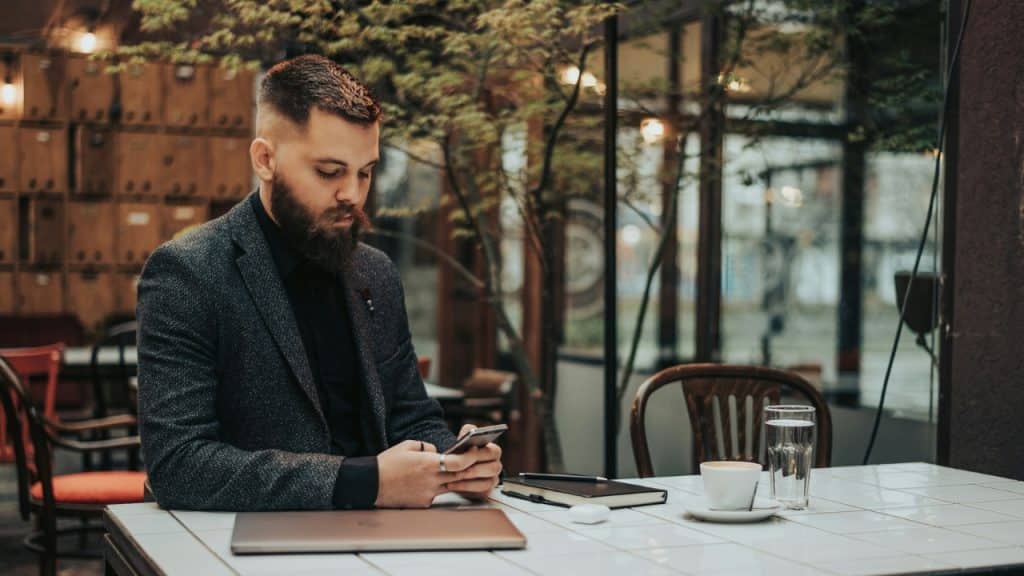 A bearded man in a blazer looks down at his phone while sitting at a table.