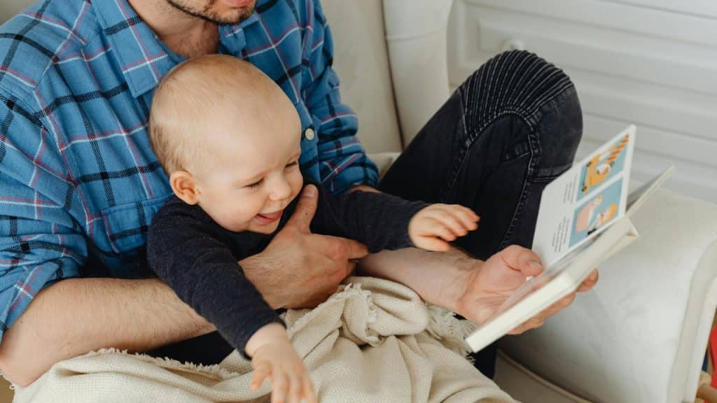 A baby sitting on an adult’s lap while looking at a book.