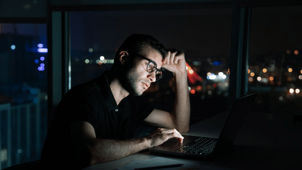 A man wearing glasses works on a laptop at night in front of a window.
