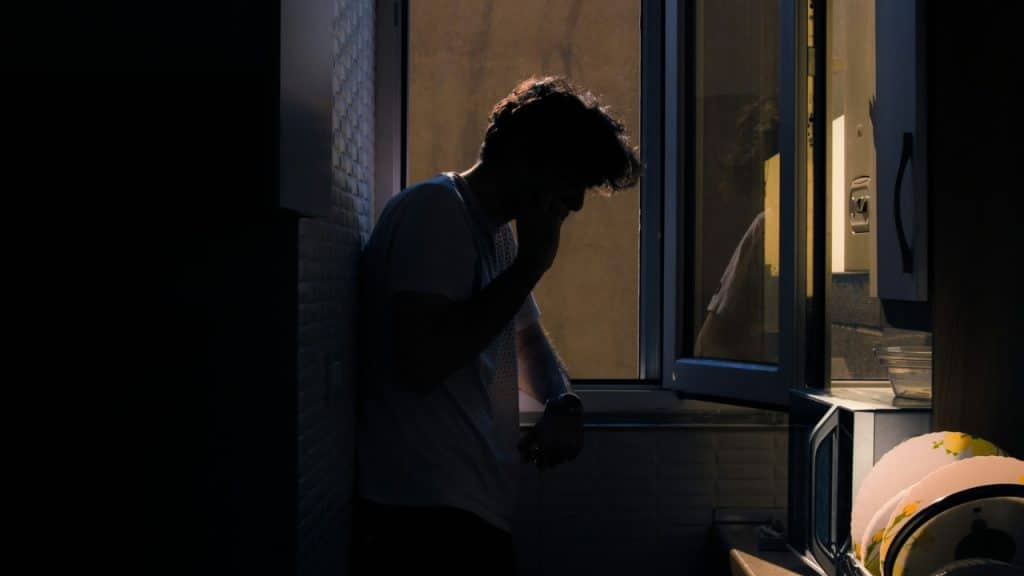 A man's silhouette stands by an open window in a dark room with dishes in the foreground.