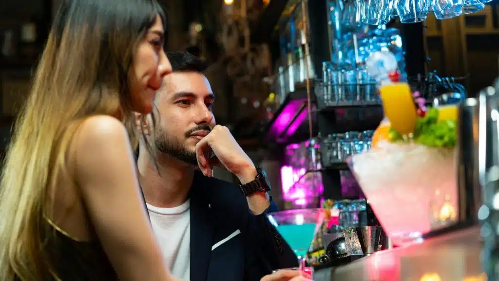 A couple sitting at a bar with colorful cocktails under neon lights.