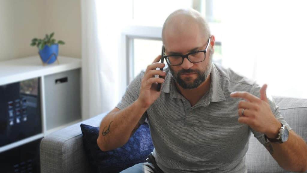 A person sitting on a couch, talking on a phone and gesturing with one hand.