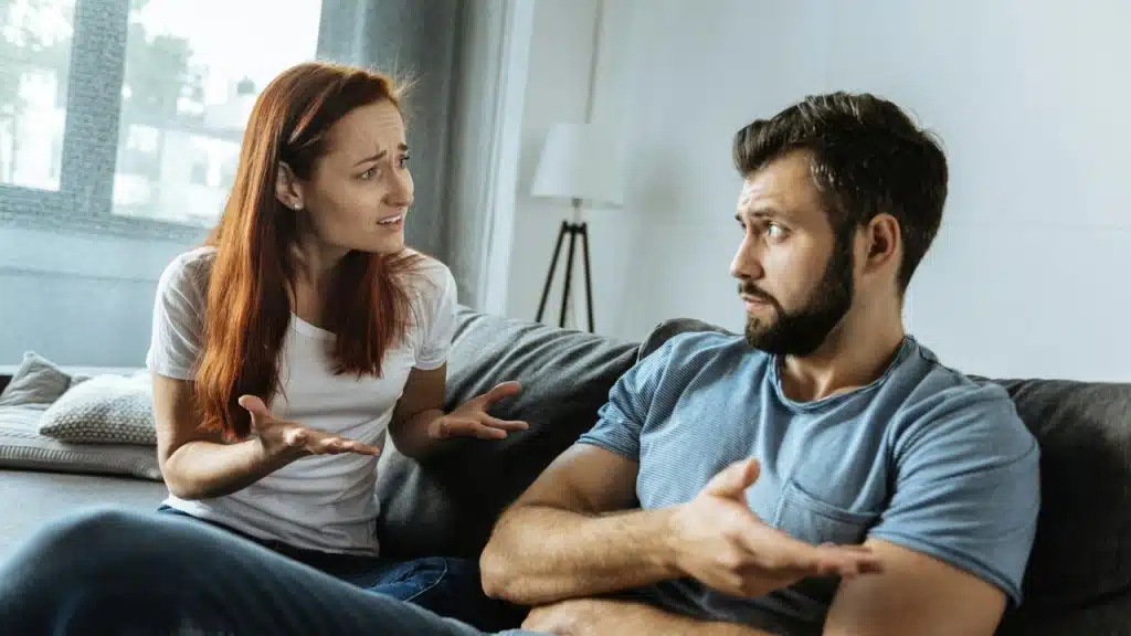A woman and man sit on a couch gesturing toward each other with concerned expressions.