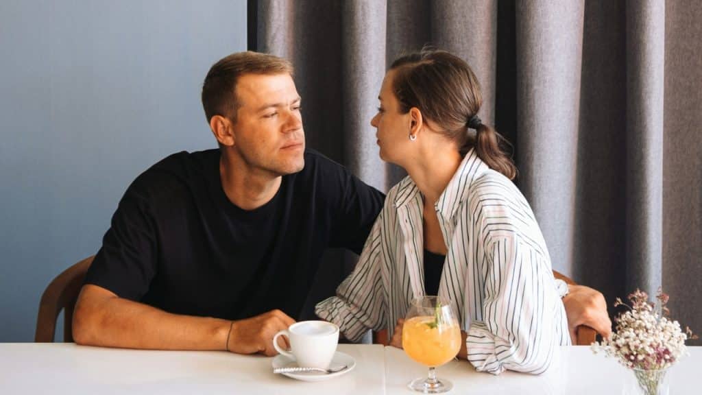 A man and woman sit at a table looking at each other with drinks present.