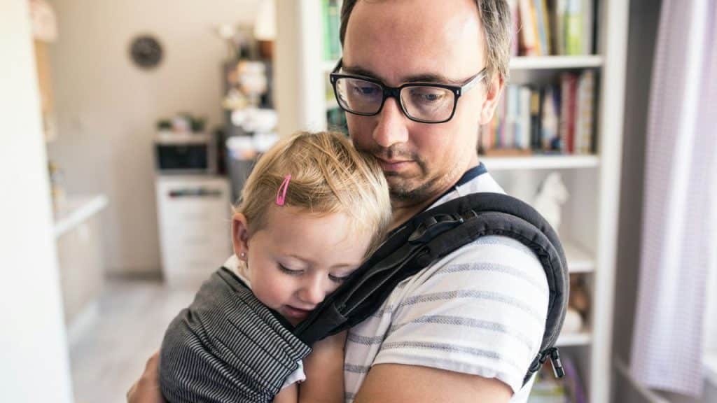 A man in glasses holds a sleeping toddler close in a front-facing baby carrier.