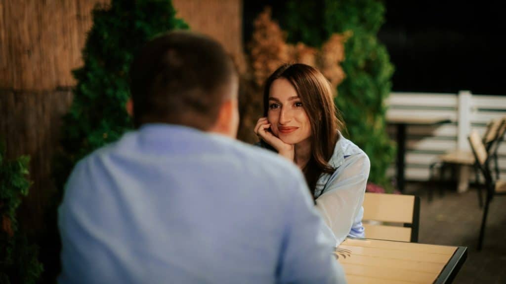 A woman rests her chin on her hand and smiles at a man sitting across.